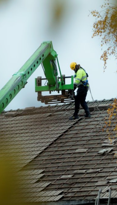 Kuvassa purettavan rakennuksen vesikatto, josta osa tiiliä purettu. Kuvassa näkyy myös rakennustyöntekijä ja vihreä nosturi, johon työntekijä lastaa kattotiiliä.